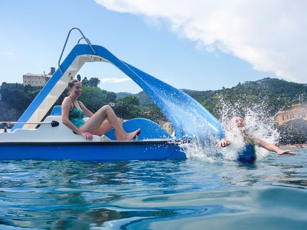 A girl splashes into the water off a floating slide attached to a boat pedalled by a smiling woman, Cinque Terre, Italy.