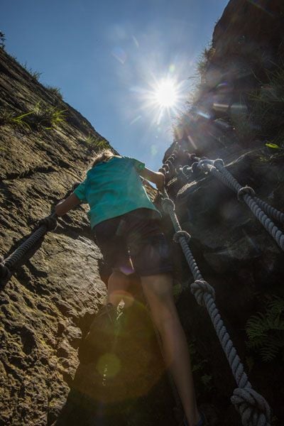 Kara scales a steep rock face using a rope ladder as the sun shines overhead in a clear blue sky.