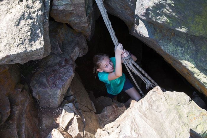 A young girl, Kara, hangs onto a rope with both hands as she pulls herself out of the top of a cave.