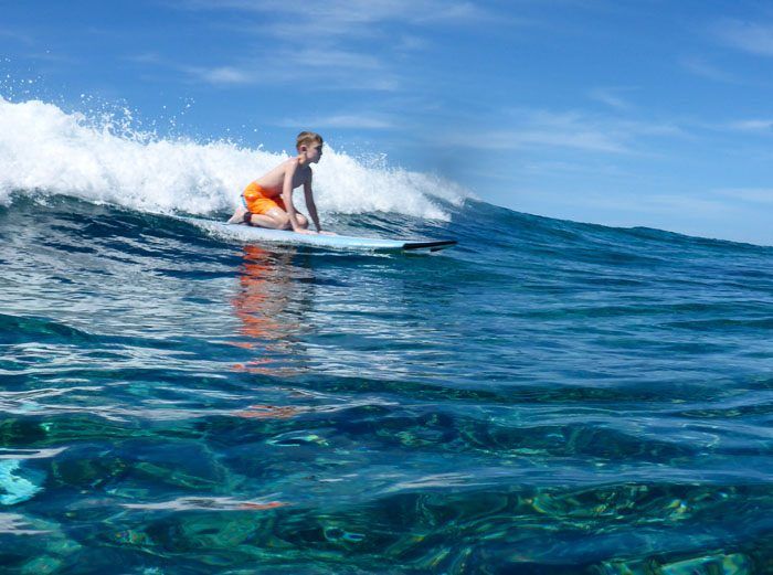 Tyler kneeling on a surfboard, surfing a wave.