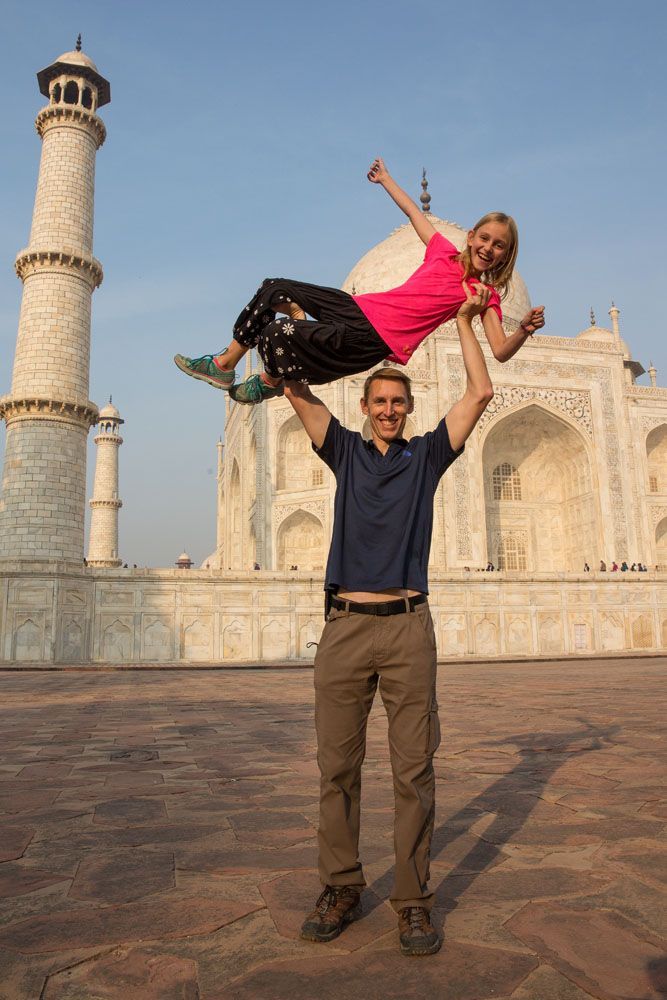 Tim holds his young daughter over his head with both arms while standing in front of the Taj Mahal. Both are smiling.
