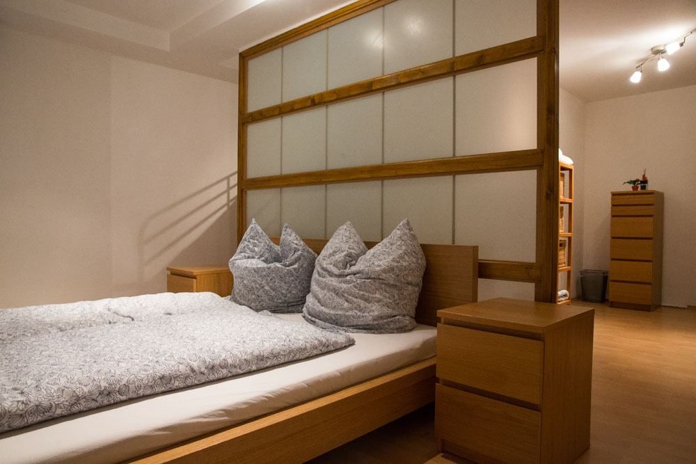 A neat bedroom with a wooden double bed base, two wooden bedside cabinets and a wooden and glass room divider for privacy.