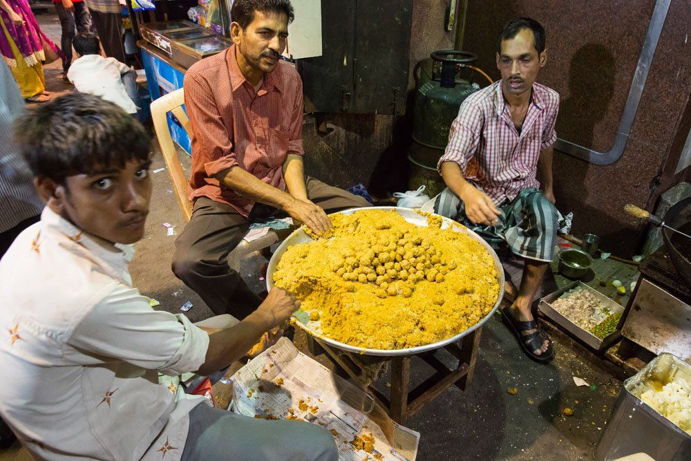 Three seated men rolling beef meatballs in a large round dish of yellow spices before cooking them.