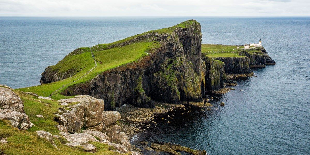 a cliff with grass and rocks by the water with Neist Point in the background