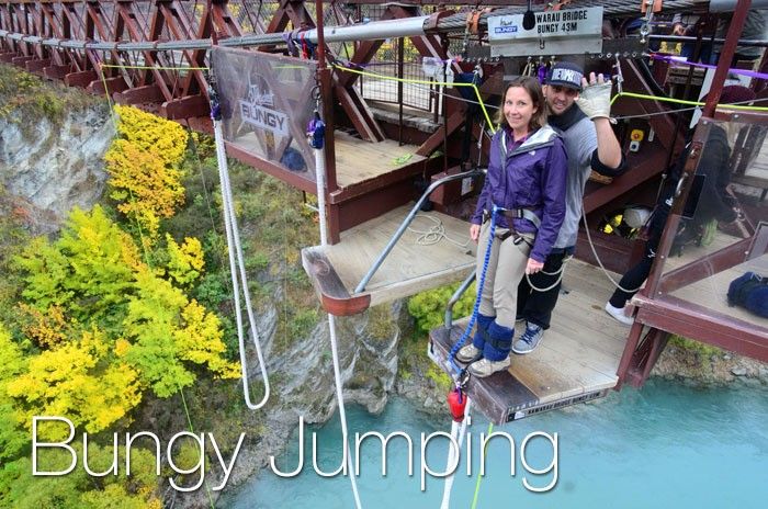 A woman in a purple jacket prepares to Bungy jump, while the guide in the back waves at the camera.
